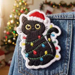 May include: An embroidered patch featuring a black cat wearing a red and white Santa hat, wrapped in a string of colorful Christmas lights, attached to blue denim fabric. The cat has large yellow eyes and is depicted in a sitting position.