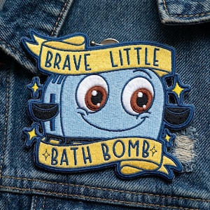 May include: Embroidered patch featuring a smiling, cartoon-style bath bomb character in light blue with brown eyes. The patch has a yellow banner that reads "BRAVE LITTLE BATH BOMB" in dark blue lettering. The patch is attached to a denim jacket.