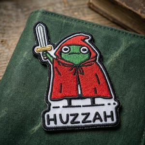 May include: Embroidered patch featuring a green frog wearing a red hooded cloak and holding a sword. The patch has a white border and the word "Huzzah" at the bottom. The patch is on a green surface.