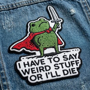 May include: Embroidered patch featuring a green frog wearing a red cape and holding a sword. The patch has the text "I HAVE TO SAY WEIRD STUFF OR I'LL DIE" in black letters on a white background. The patch is on a blue denim jacket.