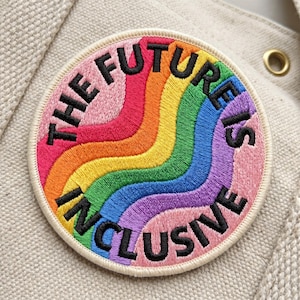 May include: Embroidered patch with a rainbow design and the words "THE FUTURE IS INCLUSIVE" in black. The patch has a cream-colored border and is set against a beige fabric background. The rainbow colors include red, orange, yellow, green, blue, and purple.