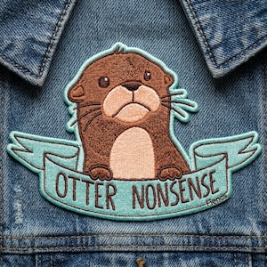 May include: Embroidered patch featuring a brown otter with a light brown belly and a sad expression. The otter is set against a light blue banner that reads "OTTER NONSENSE" in white letters. The patch is sewn onto a denim jacket.