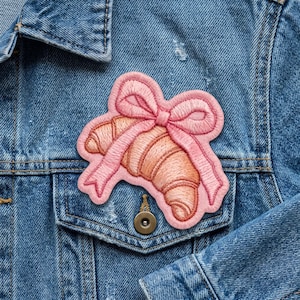 May include: Embroidered patch featuring a pink bow wrapped around a croissant. The patch has a light pink border and is sewn with detailed stitching. The patch is placed on a blue denim jacket.