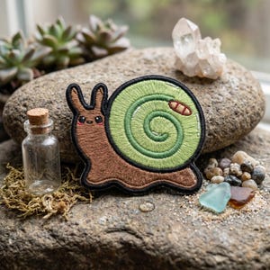 May include: Embroidered snail patch with a brown body, green shell, and black outline. The snail has a smiling face and a small red detail on its shell. The patch is on a gray rock with a small glass bottle and other decorative elements.