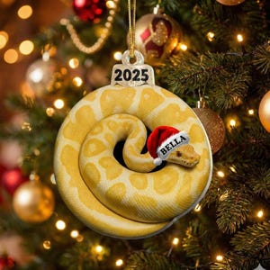 May include: A yellow and white snake Christmas ornament with a Santa hat that says "BELLA". The ornament is shaped like a coiled snake and has the year "2025" printed on the top. The ornament is hanging on a Christmas tree.
