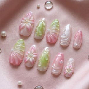 May include: A collection of ten artificial nails in various designs. The nails feature a combination of pink, white, and green colors, with pearl accents and floral or water droplet patterns. The nails are arranged on a pink surface.