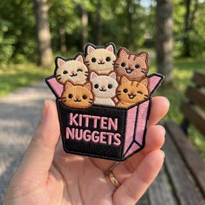 May include: Embroidered patch featuring six cartoon kittens in a pink box. The box has the words "Kitten Nuggets" in pink lettering on a black background. The kittens are various shades of tan, brown, and white.