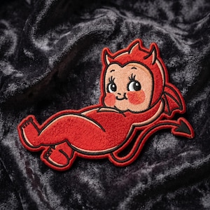 May include: Embroidered patch featuring a cartoon devil baby in red with a black outline. The devil baby has horns, wings, and a tail. The patch is on a dark, textured background.