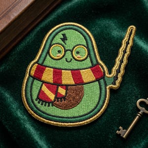May include: Embroidered patch featuring a green avocado with glasses, a scarf, and a lightning bolt scar, holding a wand. The patch has a gold border and is set against a green velvet background. A key is in the bottom right corner.