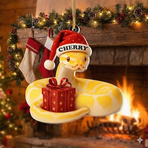 May include: A Christmas ornament featuring a yellow snake wearing a Santa hat with the word "CHERRY" on it. The snake is holding a red gift box with a red bow. The background includes a fireplace and Christmas decorations.