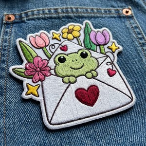 May include: Embroidered patch featuring a green frog peeking out of a white envelope. The envelope is decorated with a red heart, pink and purple tulips, yellow stars, and a pink flower. The patch is on a blue denim background.