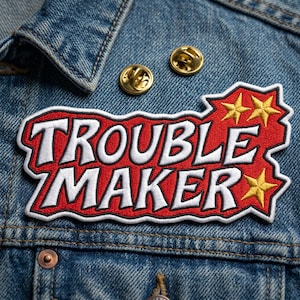 May include: A red and white embroidered patch with the words "TROUBLE MAKER" in bold, white letters. The patch has a black border and yellow star accents. Two gold-colored lapel pins are also visible on the denim jacket.