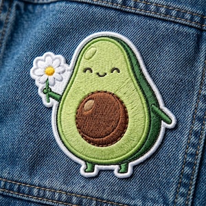 May include: Embroidered avocado patch with a smiling face, holding a white daisy. The patch features shades of green, brown, and white, with a white border. The patch is sewn onto a blue denim jacket.