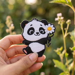 May include: Embroidered patch featuring a smiling panda bear holding a daisy. The panda has black and white fur with a pink nose and feet. The daisy has white petals and a yellow center. The patch is held in a hand.