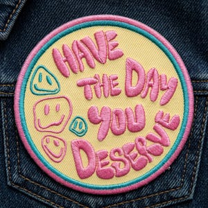 May include: Embroidered patch with the text "HAVE THE DAY YOU DESERVE" in pink, surrounded by a light yellow circle with a teal and pink border. Smiley faces are also included in the design. The patch is on a denim background.