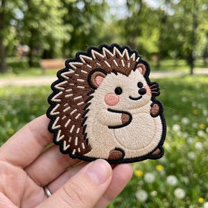 May include: Embroidered patch featuring a cartoon hedgehog. The hedgehog has a beige body, brown quills, and a black outline. It has a smiling face with pink cheeks and is holding its arms out. The patch is on a black background.