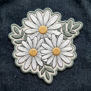 May include: Embroidered patch featuring three white daisies with yellow centers and green leaves. The patch has a scalloped edge with a light green border. The patch is on a blue denim background.