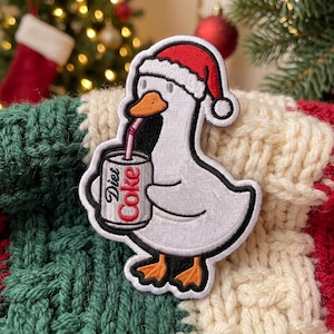 May include: An embroidered patch featuring a white duck wearing a red and white Santa hat. The duck is holding a silver can labeled Diet Coke with a pink straw. The patch has a thick black border and orange feet, set against a knitted fabric.