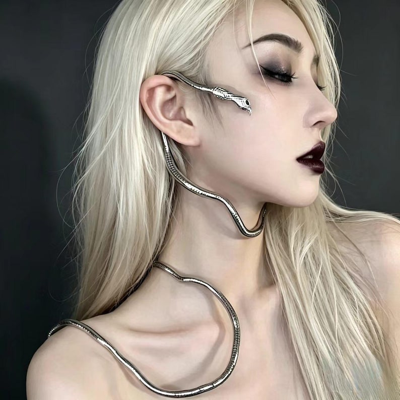 May include: A silver-toned metal ear cuff and necklace combination shaped like a long, slender snake. The snake head rests near the ear, while the elongated, segmented body curves down from the ear, wraps around the neck, and rests on the collarbone.
