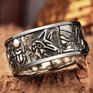 May include: A wide silver-toned metal ring featuring an oxidized finish with embossed Egyptian-inspired motifs, including a central jackal figure, a scarab beetle, and various hieroglyphic symbols engraved across the entire outer surface of the band.