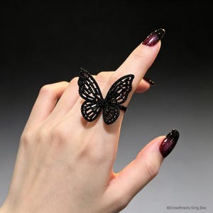 May include: A black ring featuring a large, intricate butterfly design with open-work wings encrusted with small, dark, sparkling stones. The butterfly motif is centered on the band, creating a bold, dark aesthetic with a detailed, symmetrical wing pattern.