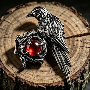 May include: A silver-toned metal ring featuring a detailed raven perched beside a sculpted rose. The center of the rose holds a smooth, round, deep red gemstone. The metalwork displays intricate feather textures and curved, organic petal shapes.