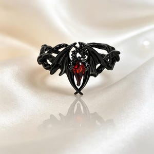 May include: A black metal ring featuring a detailed dragon motif with spread wings. The center of the dragon design is set with a faceted, diamond-shaped red gemstone. The band of the ring has a textured, vine-like appearance in black metal.