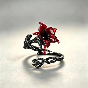 May include: A black metal ring featuring a vine-like band and a glossy red flower accent. The flower design includes a single red droplet detail extending downward from the petals, contrasting against the dark, textured, twisted metal vine.