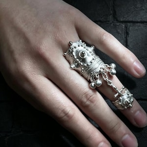 Peut inclure: Une bague argentée couvrant tout le doigt, dotée d'une base cylindrique texturée avec des clous en relief et un dôme central. La pièce comprend une chaîne reliant une bague plus petite, ornée de plusieurs petites sphères argentées pendantes.