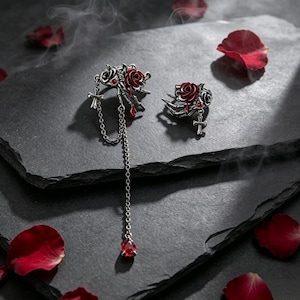May include: A pair of silver-toned ear cuffs featuring skeletal hand motifs clutching red roses. One cuff includes a dangling silver chain with a teardrop-shaped red crystal pendant and a small cross charm, while the other features a small cross.