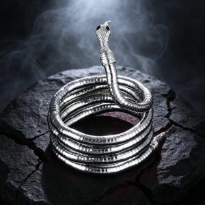 May include: A silver-toned metal snake bracelet coiled into multiple circular layers. The snake head features a textured scale pattern and two small black eyes. The body of the snake is flexible and smooth, forming a continuous spiral shape.