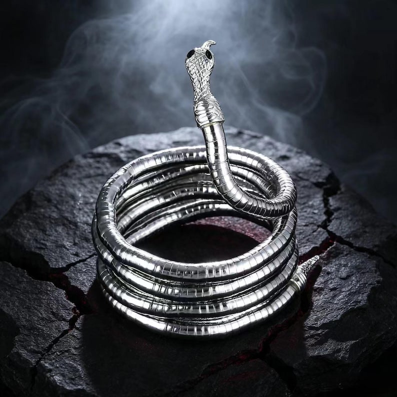 May include: A silver-toned metal snake bracelet coiled into multiple circular layers. The snake head features a textured scale pattern and two small black eyes. The body of the snake is flexible and smooth, forming a continuous spiral shape.