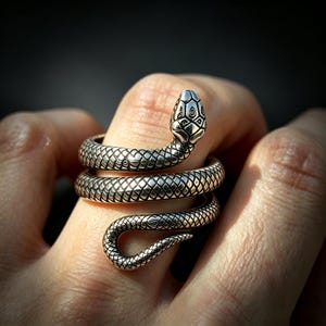 May include: A silver-toned ring shaped like a coiled snake that wraps around the finger. The metal surface features detailed, etched scale patterns across the entire body of the serpent, with a defined head and a tapered tail at the end.