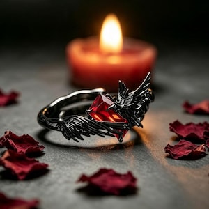 May include: A black metal ring featuring a detailed raven with outstretched wings clutching a rectangular, faceted red gemstone. The band is dark and metallic, showcasing intricate feather textures on the bird's wings and body.