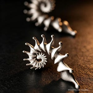 May include: Two silver-toned ear cuffs with a spiralling, ammonite-inspired shape featuring pointed, fin-like protrusions along the outer edge. The metal has a matte finish and the text Crowthrons Orig.Des is visible in the bottom right corner.