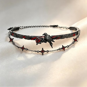 Op de afbeelding: Een zwarte metalen choker met een centraal raven- en rozenmotief met rode emaille accenten. Het ontwerp bevat een stijve band met rode stervormige uitsparingen en een delicate bungelende ketting versierd met kleine rode kristallen sterbedels.