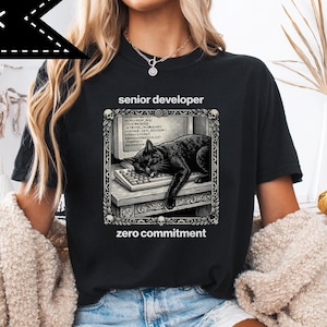 Może przedstawiać: Czarna koszulka z białym banerem z napisem "BLACK". Koszulka przedstawia grafikę czarnego kota śpiącego na klawiaturze komputera, ze słowami "senior developer" i "zero commitment".