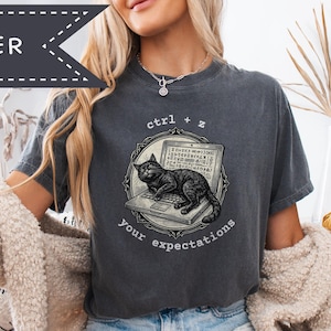 Puede incluir: Camiseta gris oscuro con un gráfico de un gato negro en un portátil, con el texto "ctrl + z your expectations". La camiseta es de corte holgado y tiene mangas cortas. La palabra "PEPPER" está impresa en una pancarta en la esquina superior izquierda.