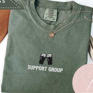 May include: Moss green t-shirt with the words "Support Group" and a graphic of two beer glasses. The shirt has a crew neck and short sleeves. A gold necklace is draped across the shirt.