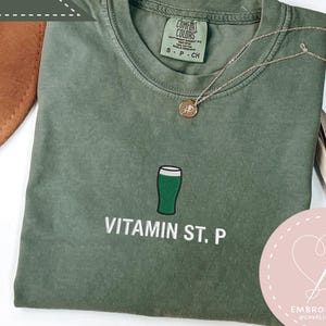 May include: A moss green t-shirt with a green beer glass graphic and the text "Vitamin St. P". The shirt has a crew neck and short sleeves. A gold necklace is draped across the shirt. A brown hat is in the background.