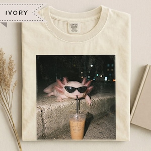 May include: An ivory-coloured t-shirt featuring a graphic print of a pink axolotl wearing black sunglasses. The axolotl is positioned behind a clear plastic cup filled with an iced coffee beverage and a black straw. The shirt has a crew neckline.