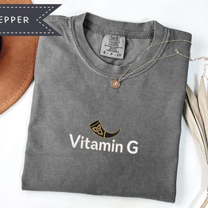 May include: A gray t-shirt with the text "Vitamin G" and a horn graphic. A gold necklace with a pendant is on the shirt. The shirt is folded and next to a brown hat and a dried plant.