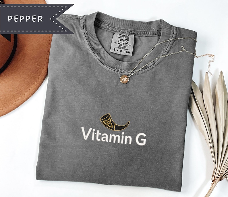 May include: A gray t-shirt with the text "Vitamin G" and a horn graphic. A gold necklace with a pendant is on the shirt. The shirt is folded and next to a brown hat and a dried plant.