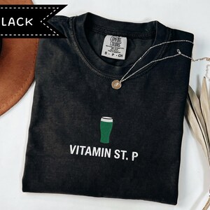 May include: Black t-shirt featuring a green beer glass graphic and the words "VITAMIN ST. P" in white. A silver necklace with a pendant rests on the shirt. The shirt is labelled "Comfort Colors" and is size S-P-CH.