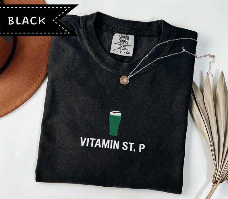 May include: Black t-shirt featuring a green beer glass graphic and the words "VITAMIN ST. P" in white. A silver necklace with a pendant rests on the shirt. The shirt is labelled "Comfort Colors" and is size S-P-CH.