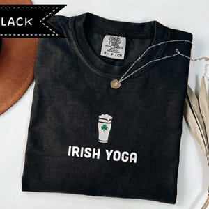 May include: Black t-shirt with the words "IRISH YOGA" in white. The shirt features a graphic of a beer glass with a shamrock. A silver necklace with a round pendant is on the shirt. The shirt is on a white surface.