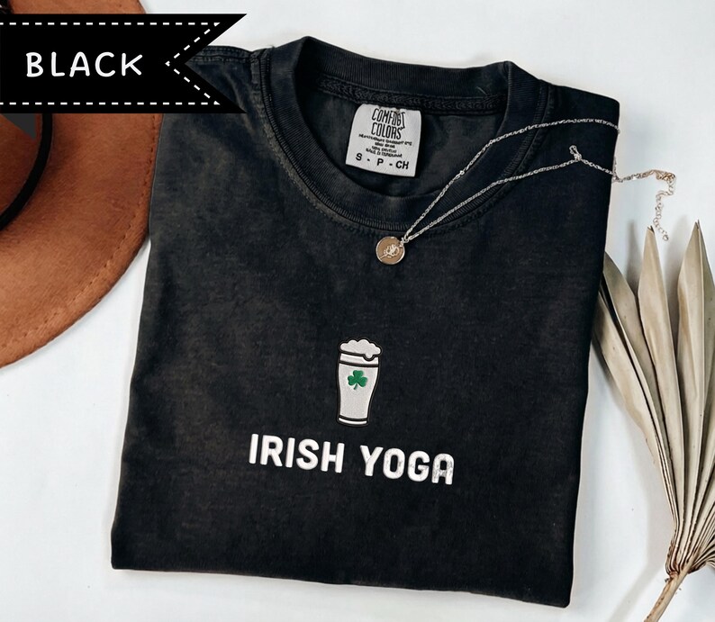 May include: Black t-shirt with the words "IRISH YOGA" in white. The shirt features a graphic of a beer glass with a shamrock. A silver necklace with a round pendant is on the shirt. The shirt is on a white surface.