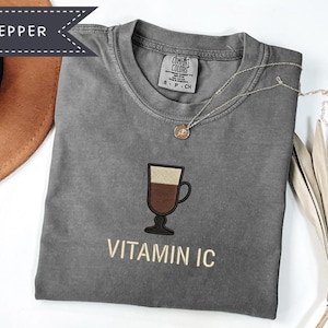 May include: A gray t-shirt with a coffee cup graphic and the text "VITAMIN IC". The shirt has a crew neck and short sleeves. A gold necklace with a pendant is draped on the shirt. A brown hat is visible in the upper left corner.