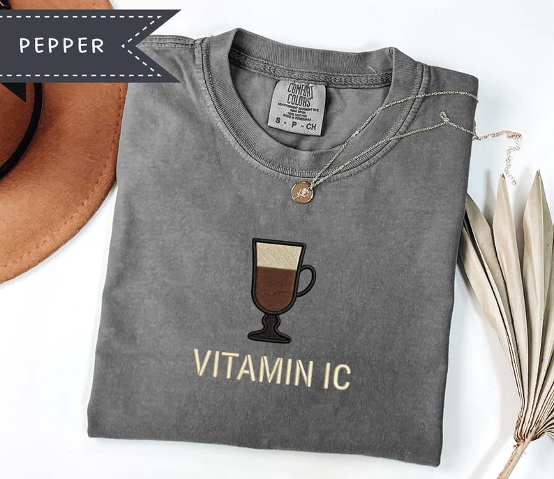 May include: A gray t-shirt with a coffee cup graphic and the text "VITAMIN IC". The shirt has a crew neck and short sleeves. A gold necklace with a pendant is draped on the shirt. A brown hat is visible in the upper left corner.