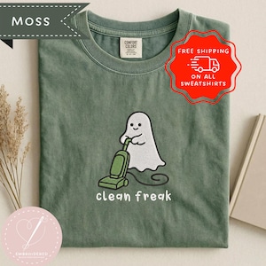 Puede incluir: Una camiseta de cuello redondo color verde musgo con un bordado de un fantasma blanco usando una aspiradora verde. Debajo del fantasma, el texto clean freak aparece en letras minúsculas blancas. La camiseta tiene una etiqueta de Comfort Colors.